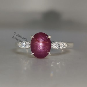 Può includere: Un anello d'argento con una pietra preziosa ovale di rubino rosso e pietre preziose più piccole trasparenti sulla fascia.