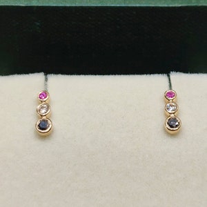 Può includere: Un paio di orecchini color oro, ciascuno con tre pietre preziose graduate: una rosa, una bianca e una blu scuro. Gli orecchini sono esposti all'interno di un portagioie verde con interno bianco.