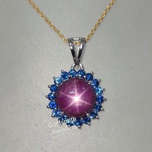 May include: A silver pendant necklace with a large, star-shaped ruby surrounded by a halo of smaller blue sapphires. The necklace is on a gold chain.
