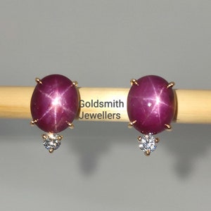 Könnte beinhalten: Ein Paar goldene Ohrringe mit einem roten Sternrubin-Edelstein und einem kleinen Diamanten-Akzent. Die Ohrringe werden auf einer Holzoberfläche mit dem Text "Goldsmith Jewellers" gezeigt.