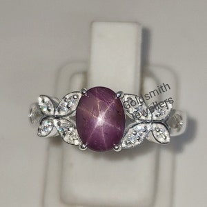 Peut inclure: Une bague en argent avec une pierre précieuse en rubis étoilé rouge et des diamants blancs sertis dans un motif floral. La bague est sur un présentoir blanc. Goldsmith Jewelers est imprimé sur le présentoir.