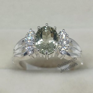 May include: A silver ring with a large oval green gemstone surrounded by smaller white gemstones. The ring is in a white box with the text "Goldsmith Jewellers".