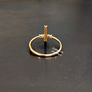 May include: Gold ring with a cross design set with small clear stones. The ring is on a black surface.