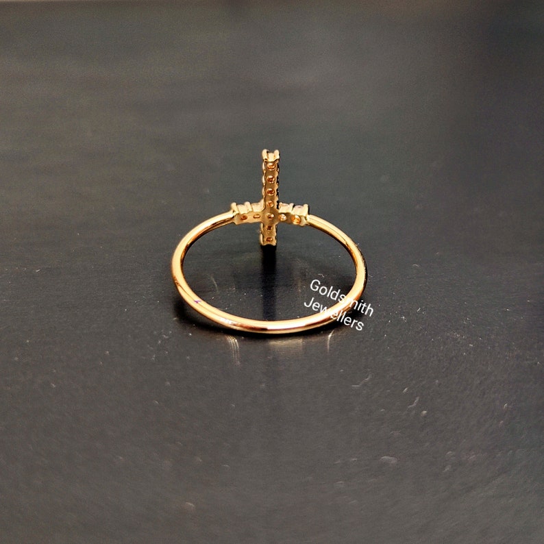 May include: Gold ring with a cross design set with small clear stones. The ring is on a black surface.