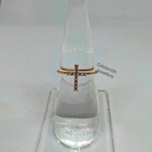 May include: A gold ring with a cross design set with clear cubic zirconia stones. The ring is displayed on a clear stand. Goldsmith Jewellers is printed on the stand.
