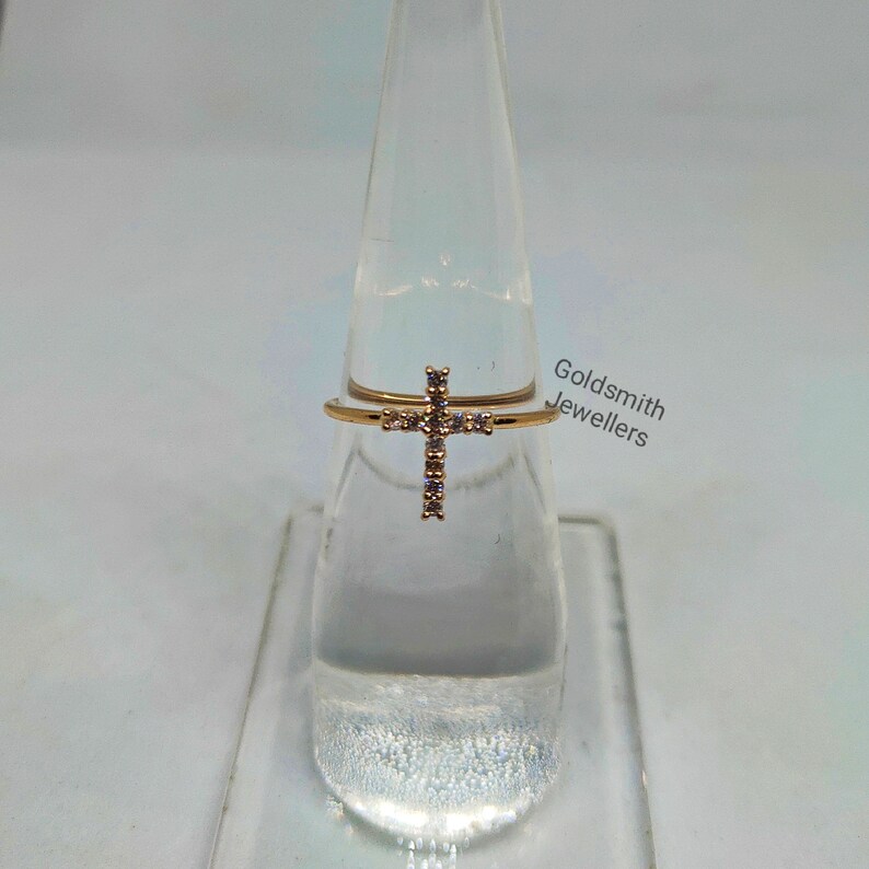 May include: A gold ring with a cross design set with clear cubic zirconia stones. The ring is displayed on a clear stand. Goldsmith Jewellers is printed on the stand.