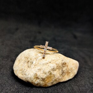 May include: A gold ring with a small, clear crystal cross design. The ring is sitting on a light brown rock.