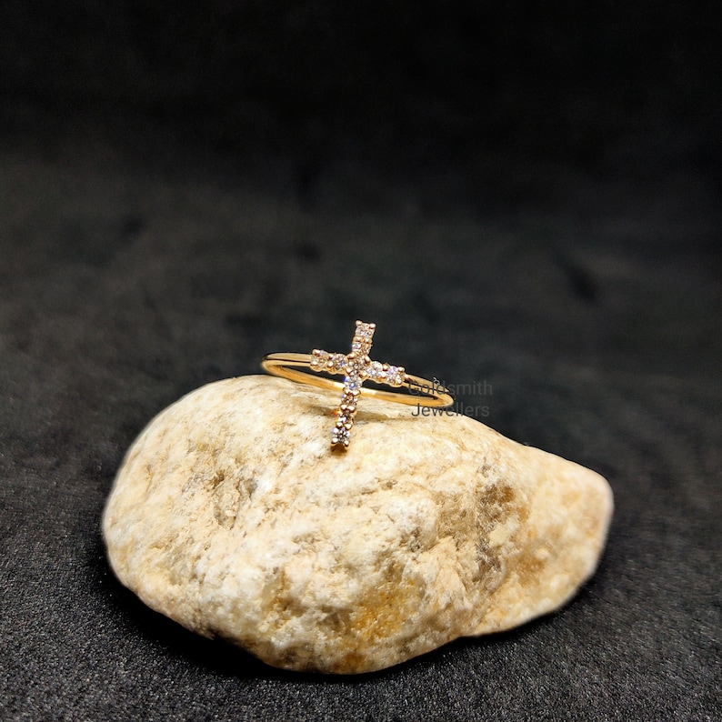 May include: A gold ring with a small, clear crystal cross design. The ring is sitting on a light brown rock.