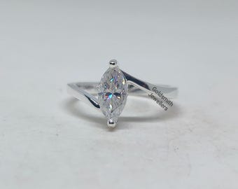 Pode incluir: Um anel de prata com uma gema de corte marquesa. A gema é o ponto focal, refletindo a luz. A banda do anel tem um design moderno e minimalista. O texto "Goldsmith Jewellers" é visível.