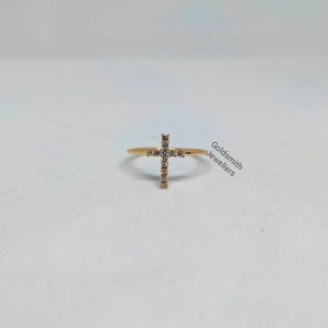 May include: A gold ring with a small cross design set with clear cubic zirconia stones. The ring is on a white background.