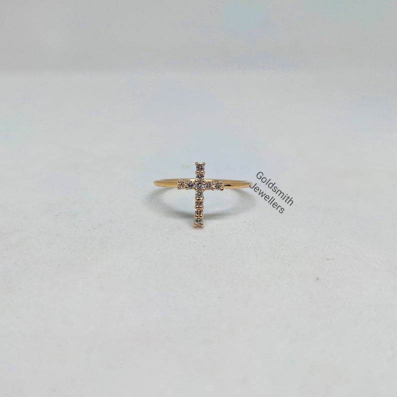 May include: A gold ring with a small cross design set with clear cubic zirconia stones. The ring is on a white background.