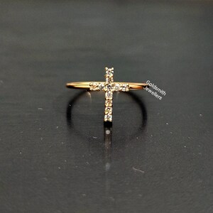 May include: A gold ring with a cross design set with small clear stones. The ring is on a black surface. Goldsmith Jewellers.