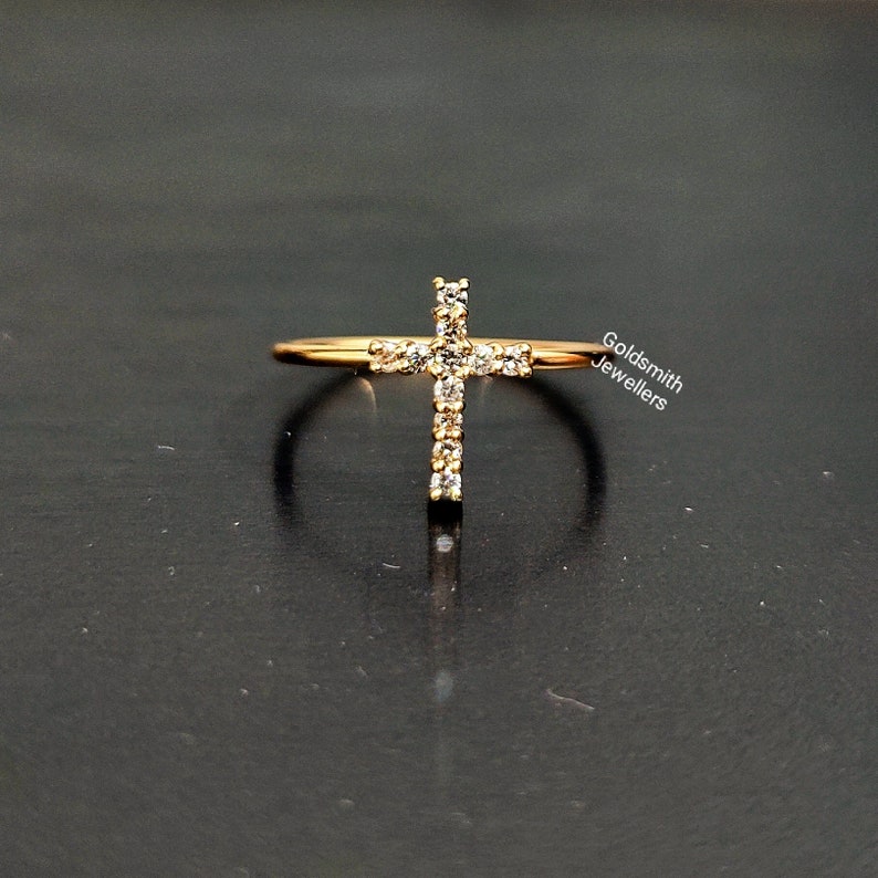 May include: A gold ring with a cross design set with small clear stones. The ring is on a black surface. Goldsmith Jewellers.