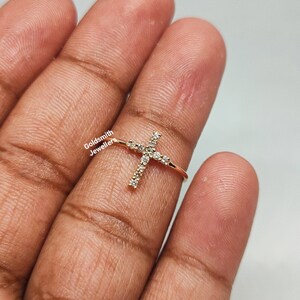May include: Gold ring with a cross design set with small clear stones. The ring is on a finger.