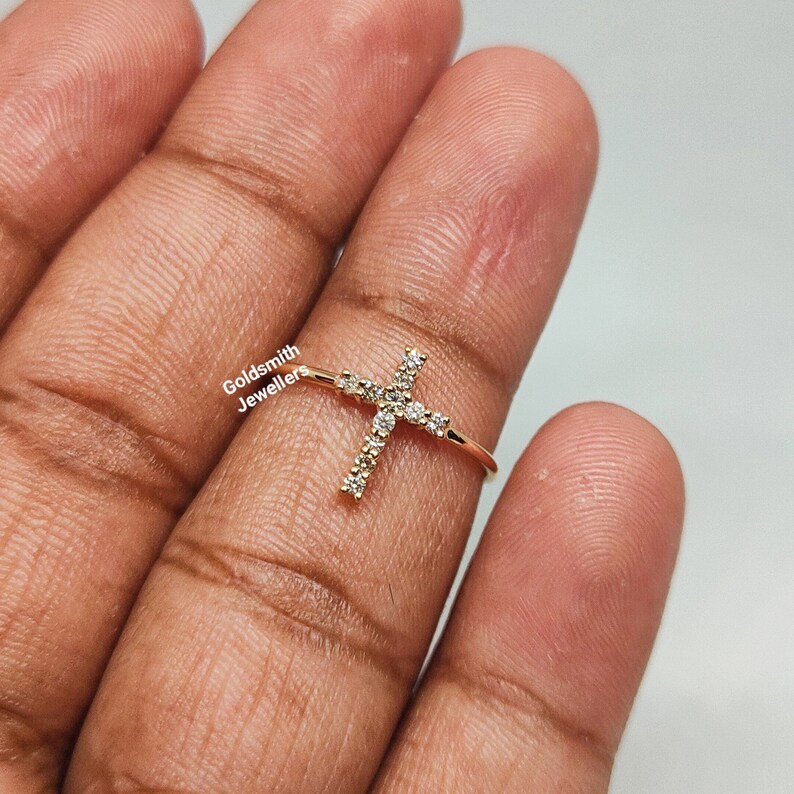May include: Gold ring with a cross design set with small clear stones. The ring is on a finger.