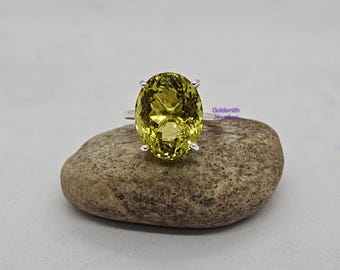 May include: A silver ring featuring a large, oval-cut, yellow-green gemstone. The gemstone is set with four prongs and sits atop a simple silver band. The ring is displayed on a textured, light brown stone against a neutral background.