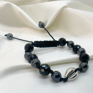 May include: Black beaded bracelet with a silver seashell charm. The bracelet is made with black beads and a black cord. The seashell charm is silver and has a detailed design.