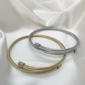 Puede incluir: Dos pulseras de cable, una dorada y otra plateada, sobre un fondo de tela blanca.