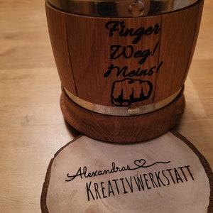 Peut inclure: Une tasse en bois avec une bande métallique et une inscription noire qui dit "Finger Weg! Meins!" avec un graphique de poing. La tasse est posée sur un sous-verre en bois avec le texte "Alexandras Kreativwerkstatt".