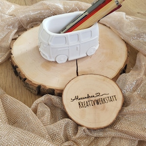 Könnte beinhalten: Ein weißer Stifthalter in Van-Form mit Stiften in verschiedenen Farben. Der Halter steht auf einer Holzscheibe, neben einem Holzuntersetzer mit dem Text "Alexandras KREATIVWERKSTATT". Der Hintergrund ist goldfarbenes Gewebe.