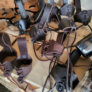 Könnte beinhalten: Eine Sammlung von braunen Leder-Becherhaltern mit Knopf und Schnürung. Die Halter hängen an einer Holzoberfläche. Der Text "ES KOMMT NICHT DRAUF AN, WAS MAN TRINKT, SONDERN MIT WEM" ist auf der Holzoberfläche sichtbar.