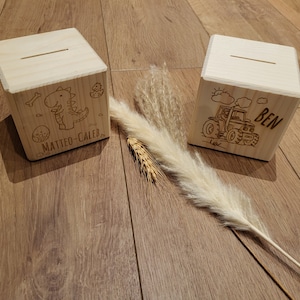 Peut inclure: Deux tirelires en bois avec des motifs gravés. L'une représente un dinosaure et le nom "MATTEO-CALEB", l'autre un tracteur et le nom "BEN". Les deux boîtes ont une fente sur le dessus pour insérer de l'argent.