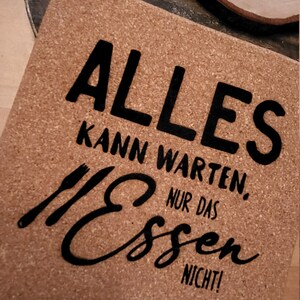 Op de afbeelding: Een kurken onderzetter met de tekst "Alles kann warten, nur das Essen nicht!" in zwarte letters. De tekst is gestileerd met een vork- en mesgrafiek.