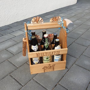 Peut inclure: Un porte-bière en bois avec une poignée et un ouvre-bouteille en métal. Le porte-bière est rempli de six bouteilles de bière. Le porte-bière est étiqueté "Werkzeugkiste" qui signifie boîte à outils en allemand.