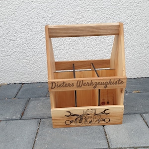 Peut inclure: Un porte-outils en bois avec une poignée, avec le texte gravé "Dieters Werkzeugkiste". L'avant du porte-outils affiche une image d'un tracteur et de clés. Le porte-outils est en bois clair et possède des séparateurs internes.