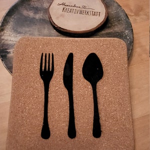 Op de afbeelding: Kurk onderzetter met een zwart silhouet van een vork, mes en lepel. De onderzetter is bruin en heeft een ruwe textuur. De tekst "Alexandras Kreativwerkstatt" staat op een ronde houten schijf.