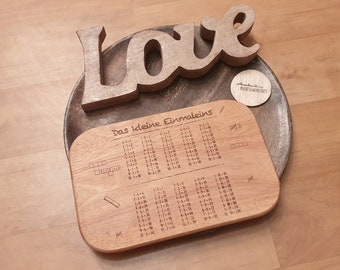 Può includere: Un tagliere di legno con le tabelline di moltiplicazione da 1 a 10 incise su di esso. Il tagliere è su una superficie di legno con un cartello di legno "Love" e un'etichetta rotonda di legno con il testo "Kreativ Werkstatt" su di esso.