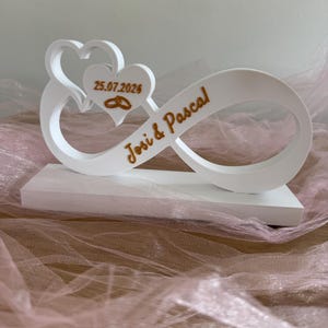 Può includere: Una scultura bianca a forma di simbolo dell'infinito su base rettangolare, con due cuori ritagliati, la data 25.07.2026, l'icona di due anelli intrecciati e la scritta dorata Josi & Pascal in rilievo sulla parte anteriore della struttura.
