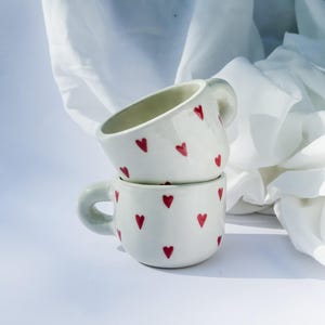 Op de afbeelding: Twee witte keramische mokken, elk versierd met kleine, rode hartvormige ontwerpen. De mokken hebben afgeronde lichamen en kleine handvatten. De achtergrond is een wit oppervlak met een witte doek.