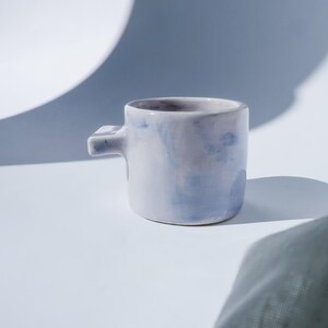 Könnte beinhalten: Eine kleine, handgefertigte Keramiktasse mit einem einzigartigen Griff. Die Tasse hat eine blasslavendelfarbene Farbe mit dezenten, aquarellartigen blauen Akzenten. Die Tasse steht auf einer weißen Oberfläche mit weichen Schatten.