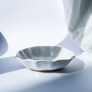 Könnte beinhalten: Eine kleine, hellgraue Keramikschale mit gewelltem Rand. Die Schale steht vor einem weißen Hintergrund mit weichen Schatten, was eine minimalistische Ästhetik suggeriert. Das Design ist schlicht und elegant, geeignet zum Servieren kleiner Portionen.