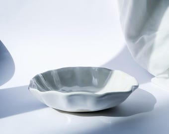 May include: A small, light gray ceramic bowl with a wavy rim. The bowl is set against a white background with soft shadows, suggesting a minimalist aesthetic. The bowl's design is simple and elegant, suitable for serving small portions.