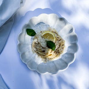 Könnte beinhalten: Draufsicht auf einen weißen Teller mit gewelltem Rand, der eine Portion Pasta enthält. Die Pasta ist mit geriebenem Käse, einer Zitronenscheibe und frischen Basilikumblättern belegt. Der Teller steht auf einer weißen Oberfläche.