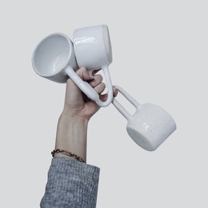 Könnte beinhalten: Drei weiße Keramikbecher mit einzigartigen Griffen werden vor einem hellgrauen Hintergrund präsentiert. Die Becher haben eine gesprenkelte Textur und ein modernes, minimalistisches Aussehen. Die Griffe sind gebogen und umschließen den Becherkörper.
