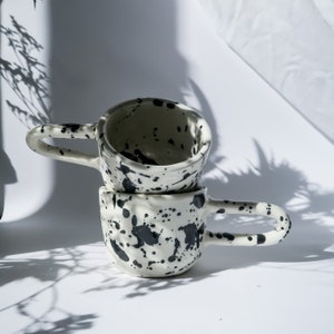 Könnte beinhalten: Zwei gestapelte weiße Keramikbecher mit schwarzem Farbspritzer-Design. Jeder Becher hat einen großen, gebogenen Henkel. Die Becher stehen auf einer weißen Oberfläche, mit Schatten von Blättern im Hintergrund.