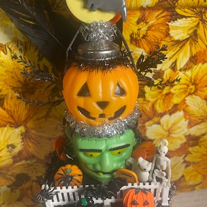 Puede incluir: Una decoración de Halloween con una figura verde de Frankenstein con cabeza de calabaza, rodeada de calabazas en miniatura, un gato negro, un esqueleto y una valla blanca. La figura está coronada con una calabaza, un murciélago y una luna.