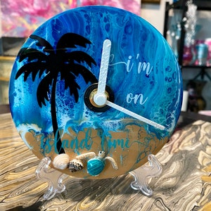 May include: A round clock with a beach theme. The clock face features a blue and white ocean design, a black palm tree silhouette, and the words "I'm on" and "Island Time". The clock is supported by a clear stand.