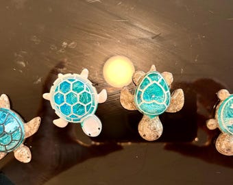 May include: Four turtle-shaped decorative ornaments featuring textured, speckled tan bodies and vibrant, swirling blue resin shells. Each shell displays a unique pattern, including geometric hexagons, a heart shape, and small star accents in white.