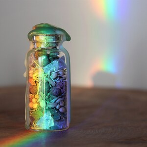 May include: A glass jar filled with colorful potpourri, sealed with a green wax lid. A rainbow of light shines on the jar, casting a colorful glow.