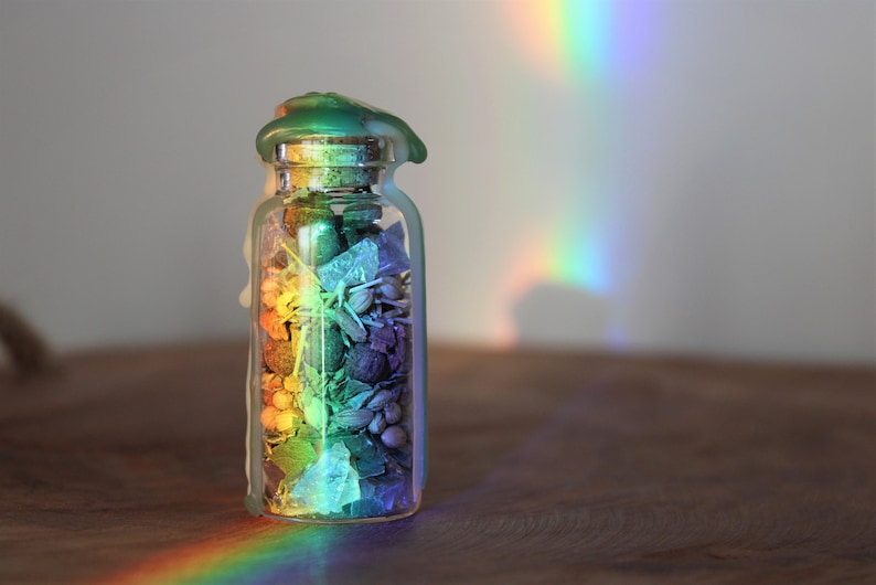 May include: A glass jar filled with colorful potpourri, sealed with a green wax lid. A rainbow of light shines on the jar, casting a colorful glow.