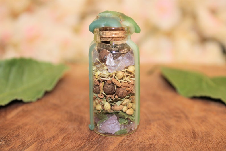 May include: A small glass bottle with a green wax seal on the top. The bottle is filled with various herbs, spices, and crystals.