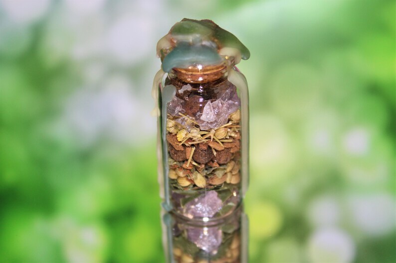 May include: A small glass jar with a cork top is filled with various herbs, spices, and crystals. The jar is sealed with a layer of melted wax, giving it a rustic and magical appearance.