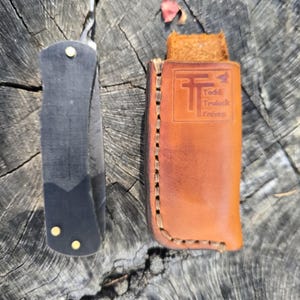 May include: A folding knife with a matte black handle secured by three brass rivets, positioned next to a tan leather sheath. The sheath features a stamped logo with the text Todd Trulock Knives and is constructed with visible stitching.