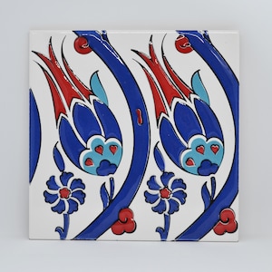 Puede incluir: Azulejo cerámico cuadrado con un patrón floral repetitivo. El diseño presenta flores azules en forma de tulipán con detalles rojos, tallos azules y pequeños detalles florales azules y rojos sobre un fondo blanco. El azulejo es probablemente para uso decorativo.