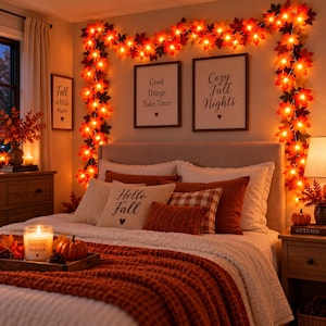 May include: A bedroom decorated with a string of glowing maple leaf lights draped over the headboard. The wall features framed prints with the text Good Things Take Time and Cozy Fall Nights. The bed has pillows with the text Hello Fall.