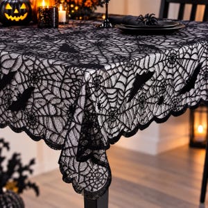 May include: A black lace tablecloth featuring an intricate spider web pattern with silhouettes of flying bats and small spiders integrated into the design. The fabric has a scalloped edge and a semi-sheer texture draped over a rectangular table.